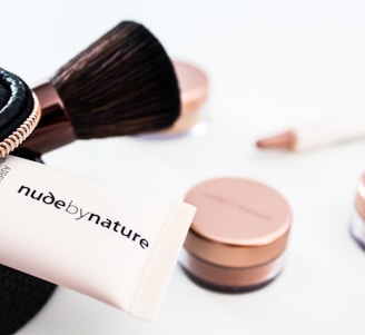 A collection of cosmetics is neatly displayed, including a makeup brush, a black zippered pouch, and several rose-gold capped jars. A tube that reads 'nude by nature' is partially visible, suggesting a beauty product brand.