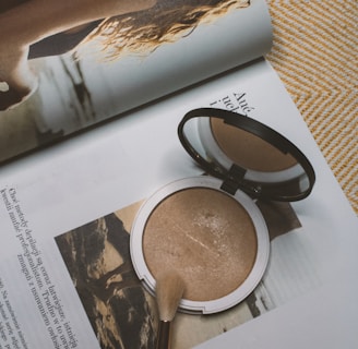 A makeup compact and brush rest on the pages of an open magazine. The magazine features an image of a woman with wavy hair looking back over her shoulder at the camera. Text is visible on the pages, and the overall setting creates a sense of everyday beauty and style.